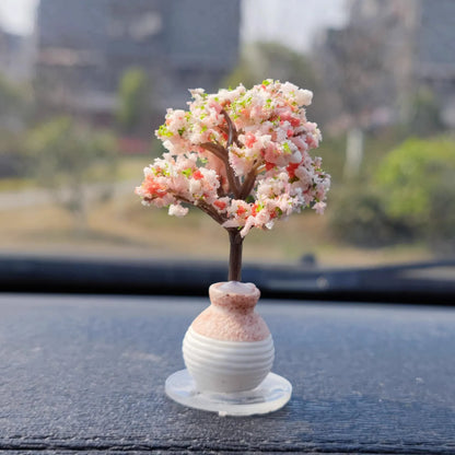 Decorative miniature tree in a white vase on a car dashboard with a blurred outdoor background