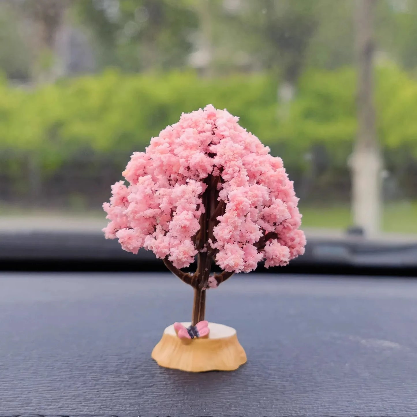 Pink cherry blossom tree figurine on a car dashboard with a blurred green outdoor background