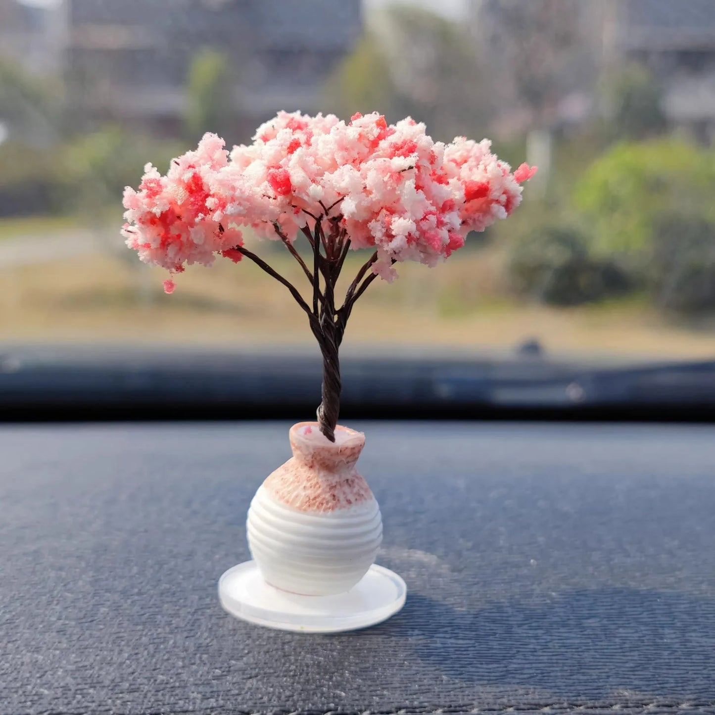 Small decorative tree with pink blossoms in a white vase on a car dashboard.