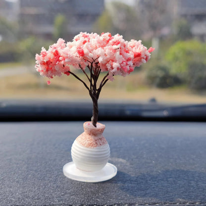 Small decorative tree with pink blossoms in a white vase on a car dashboard.