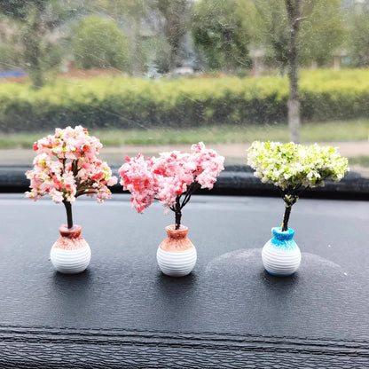 Three small decorative trees in vases on a car dashboard with a blurred outdoor background.